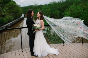 newlyweds smile together overlooking water with bride's veil floating behind them