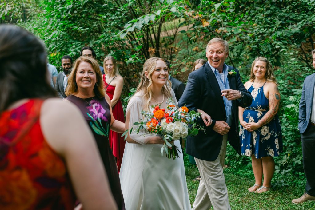 Bride is escorted by parents up the aisle during ceremony, surrounded by guests, outside